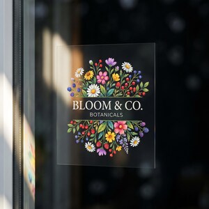 以下が含まれることがあります： 様々な花、ベリー、緑の葉が円形に配置されたカラフルな植物イラストが描かれた、正方形の透明なウィンドウデカール。デザインの中央には、白い文字で「BLOOM & CO. BOTANICALS」と印刷されています。