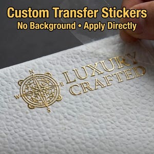 以下が含まれることがあります： 金箔転写ステッカー。コンパスローズのアイコンと「LUXURY CRAFTED」の文字が白いテクスチャーのある表面に貼られています。ステッカーは透明な台紙から剥がされています。上部には「Custom Transfer Stickers」という文字があります。