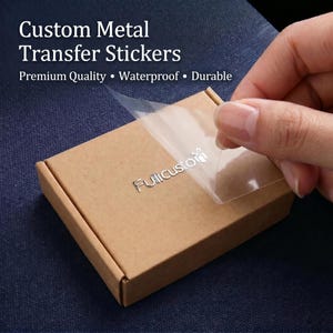 Puede incluir: Una pegatina de transferencia de metal plateado con el texto "Fullicustom" aplicada sobre una caja cuadrada de cartón marrón. Se está despegando una película adhesiva transparente de la superficie, revelando las letras metálicas.