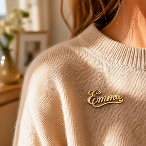 May include: A gold brooch spelling "Emma" in a cursive font. The brooch is attached to a beige jumper. The brooch is a personalized accessory.