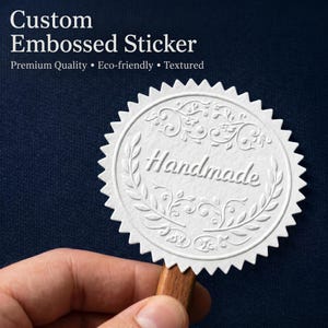 以下が含まれることがあります： 縁がギザギザになった白い円形のステッカーで、中央に「Handmade」という文字がエンボス加工され、その周りを装飾的な葉のリースとスクロール模様が囲んでいます。ステッカーの上部には「Custom Embossed Sticker Premium Quality Eco-friendly Textured」というテキストが表示されています。