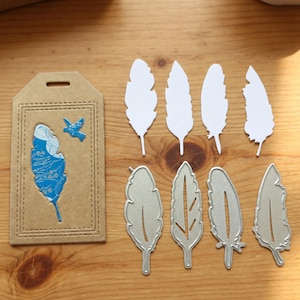 Peut inclure: Divers éléments de loisirs créatifs sur le thème des plumes. L'image présente une étiquette marron avec un motif de plume bleu et un oiseau, des plumes en papier blanc et des découpes de plumes en métal argenté.