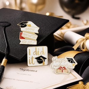 以下が含まれることがあります： 卒業をテーマにしたエナメルピン、ゴールドのアクセント付き。「Happy Graduation, You Did It」と書かれたピンが重ねられたバナーに。「I did it」と卒業帽子のピンも。卒業証書も見える。