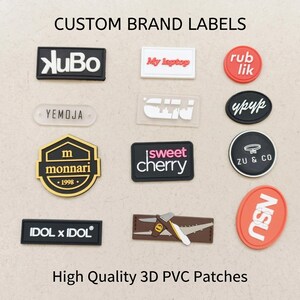 Puede incluir: Una colecci&oacute;n de etiquetas y parches de marca personalizados en PVC 3D de diversas formas, colores y tama&ntilde;os. Las etiquetas presentan varios logotipos y textos, incluyendo KuBo, My laptop, rub lik, YEMOJA, monnari 1998, sweet cherry, IDOL x IDOL, ZU & CO y NSU.