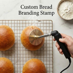 Può includere: Un timbro personalizzato per la marchiatura del pane viene utilizzato su un panino dorato. Il timbro è in ottone ed è attaccato a un cannello nero e argento. Il testo "Custom Bread Branding Stamp" è nella parte superiore dell'immagine. Una ciotola di farina è sullo sfondo.