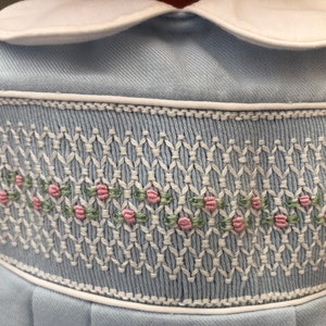 May include: A light blue baby dress with a white collar and a white embroidered trim. The trim features a repeating pattern of pink flowers and green leaves.