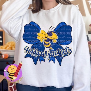 Könnte beinhalten: Weißes Sweatshirt mit einer großen blauen Schleife und einer gelb-schwarzen Cartoon-Biene in der Mitte. Darunter steht in Schreibschrift das Wort "Yellowjackets". Ein Fanartikel.