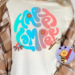 May include: A white t-shirt with a colorful graphic that says "Happy Camper" in a retro font. The graphic includes a campfire, a marshmallow on a stick, and a flower.