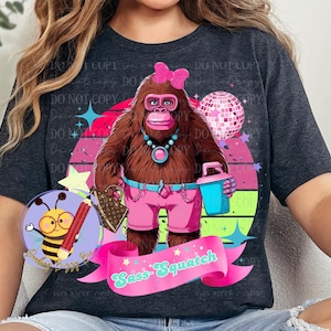 May include: Dark gray t-shirt featuring a cartoon Sasquatch character with a pink bow, shorts, and accessories. The design includes a disco ball, a pink banner with the text "Sass-Squatch", and a bee logo.