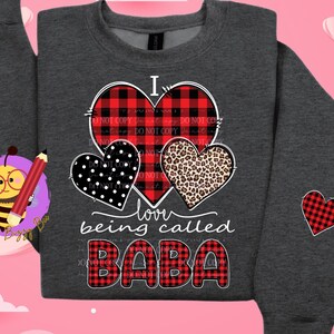May include: Dark gray sweatshirt with a design featuring three hearts: a red and black plaid heart, a black polka dot heart, and a leopard print heart. The text reads "I love being called BABA". A red plaid heart is on the sleeve.