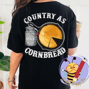 Puede incluir: Camiseta negra con el texto "Country as Cornbread" en letras blancas. El diseño presenta una sartén de pan de maíz, una jarra de leche y una cinta roja y blanca.