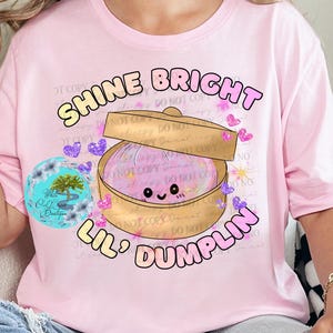 May include: A light pink t-shirt with the text "SHINE BRIGHT LIL' DUMPLIN" in a playful font. The graphic features a cartoon dumpling in a bamboo steamer, surrounded by hearts and butterflies. The shirt is a casual style.