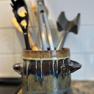 May include: A handmade ceramic utensil holder with a brown, blue, and tan glaze. The stoneware crock has two handles and holds various kitchen tools, including a whisk, tongs, and a slotted spoon. The glaze has a dripped effect.