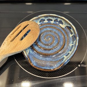 May include: A round ceramic spoon rest featuring a blue and brown spiral pattern with a reactive glaze finish. A light-colored wooden slotted spoon rests partially on top of the ceramic dish, which has a raised edge and a textured surface.