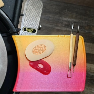 May include: A pottery wheel with a cutting board and tools. The board has a yellow, orange, and pink gradient. Tools include a wooden handled rib and a needle tool. A sponge and a red trimming tool are also present.
