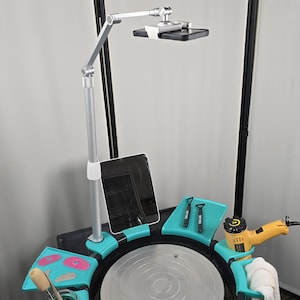 Könnte beinhalten: Ein Töpferscheiben-Setup mit einem silbernen, verstellbaren Arm, der ein Telefon und ein Tablet hält. Das Rad hat eine schwarze Basis mit türkisfarbenen Ablagen für Werkzeuge. Ein gelbes Elektrowerkzeug und ein weißes Handtuch sind ebenfalls sichtbar.