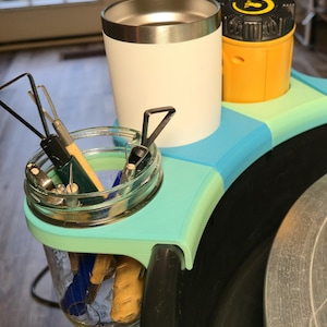 May include: A pottery wheel accessory featuring a white tumbler, a yellow container, and a glass jar filled with tools. The accessory is teal and blue, designed to hold tools and a beverage while working on the pottery wheel.