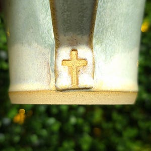 May include: Close-up of a handmade ceramic mug with a light blue and cream glaze. The mug features a gold-colored cross emblem on the handle base. The base of the mug is a natural tan color. The mug is set against a blurred green background.