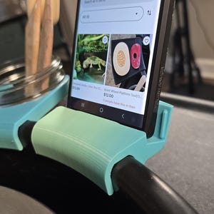 Puede incluir: Un teléfono inteligente se sostiene en un soporte impreso en 3D de color verde azulado claro, que muestra una tienda de Etsy. La tienda vende artículos como una caja de arena para gecko leopardo y una herramienta de plataforma de rueda Brent. Un frasco con lápices y una esponja está en el fondo.