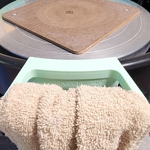 Puede incluir: Un torno de alfarero con un cuadrado de madera encima. Una bandeja verde claro con el texto "Hannah's Meadow Pottery" está frente a una toalla beige. La rueda es negra y plateada.