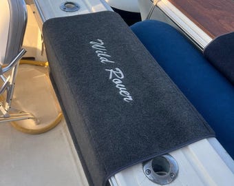 May include: A dark gray boat cushion cover with the text "Wild Rover" embroidered in white. The cover is placed on a white boat surface, with a blue seat visible in the background. A wooden boat trim is also visible.