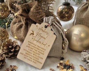Könnte beinhalten: Ein helles Holz-Geschenkanhänger mit der Gravur 'Archie's Magical Reindeer food' und Anweisungen für Heiligabend. Der Anhänger ist an einem kleinen beigen Jutebeutel mit Kordelzug befestigt, umgeben von Tannenzapfen und goldenen Ornamenten.