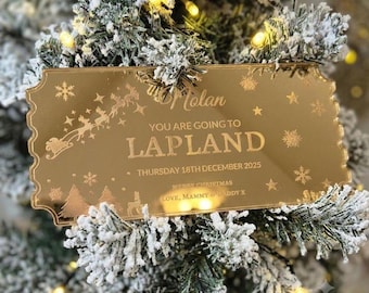 May include: A gold-toned, rectangular acrylic ticket ornament with scalloped edges, featuring engraved text that reads: Molan, YOU ARE GOING TO LAPLAND, THURSDAY 18TH DECEMBER 2025, MERRY CHRISTMAS, LOVE, MAMMY & DADDY X, surrounded by reindeer, stars, and snowflakes.