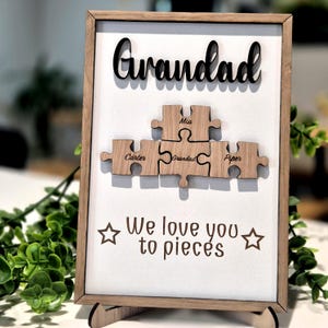 May include: Wooden framed sign with the word "Grandad" in black script at the top. Below, five wooden puzzle pieces are arranged, each with a name: Carter, Grandad, Mia, and Piper. The phrase "We love you to pieces" is printed below the puzzle pieces.