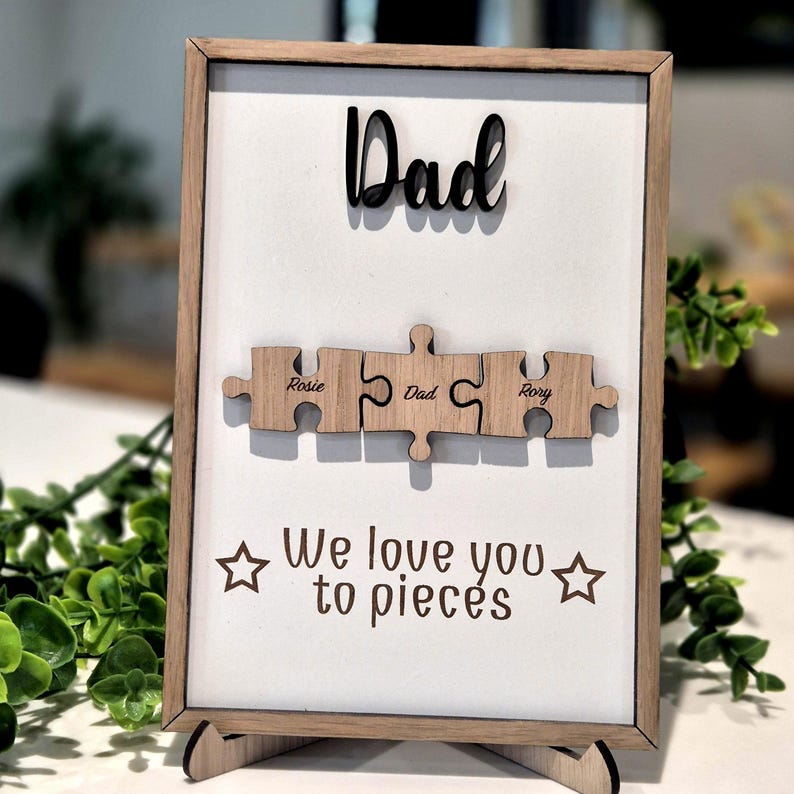 May include: A framed wooden sign with the word "Dad" in black script above three wooden puzzle pieces. The puzzle pieces are engraved with names and the word "Dad". The phrase "We love you to pieces" is printed below.
