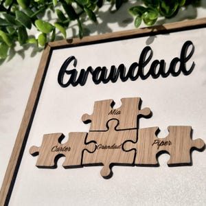 May include: A wooden framed sign with the word "Grandad" in black script above four wooden puzzle pieces. Each puzzle piece has a name engraved on it: Carter, Mia, Grandad, and Piper. The frame is light brown.