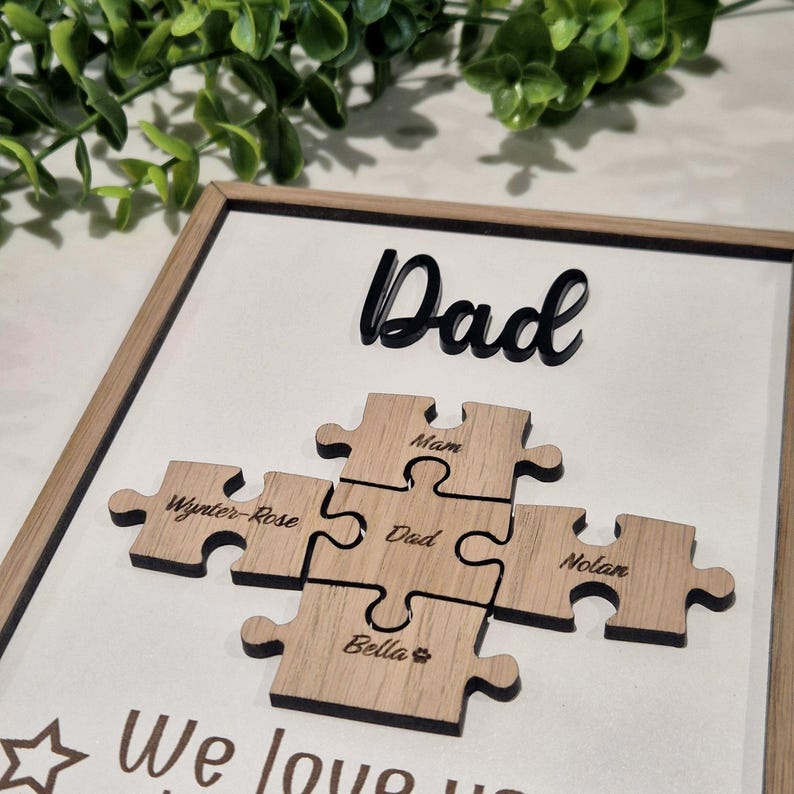 May include: A framed wooden puzzle art piece with the word "Dad" in black script. Five wooden puzzle pieces are engraved with names. The words "We love you" are visible at the bottom. A decorative gift.