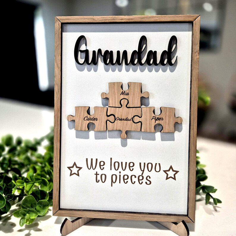 May include: Wooden framed sign with the word "Grandad" in black script above wooden puzzle pieces. The puzzle pieces have names on them. The phrase "We love you to pieces" is below the puzzle pieces.