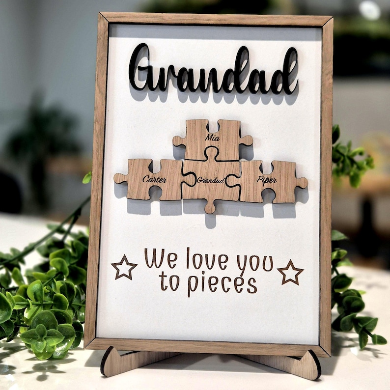 May include: Wooden framed sign with the word "Grandad" in black script above wooden puzzle pieces. The puzzle pieces have names on them. The sign reads "We love you to pieces" with star accents.