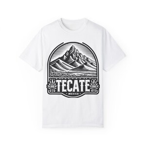 May include: White t-shirt featuring a black and white graphic of a mountain range within a circular frame. The word "TECATE" is prominently displayed below the mountains, with "MEXICO" underneath.