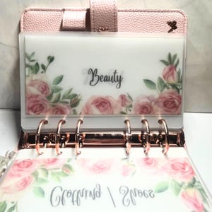 Könnte beinhalten: Rosa Kunstleder-Ordner mit roségoldfarbenen Ringen und einem Schnappverschluss. Die transparente Tasche ist mit rosa Rosen und dem Wort "Beauty" verziert. Der Ordner ist für die Organisation konzipiert und hat ein florales Thema.