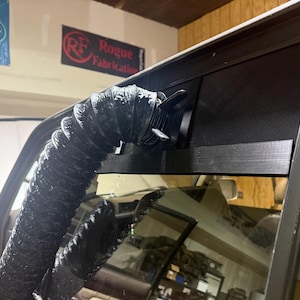 Puede incluir: Una manguera flexible negra está conectada a una ventilación en una ventana de vehículo. La ventilación está unida a una pantalla de malla negra. El texto "RF Rogue Fabrication" es visible en un letrero en el fondo.