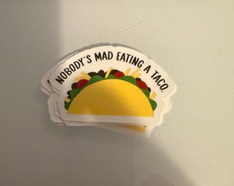 May include: A sticker featuring a cartoon taco with yellow, green, and red fillings. The text "NOBODY'S MAD EATING A TACO" is printed above the taco in a semi-circular shape. The sticker has a white border.