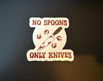 以下が含まれることがあります： 「No Spoons Only Knives」という赤い文字が書かれた白いステッカー。ステッカーの中央には血まみれのナイフが描かれている。