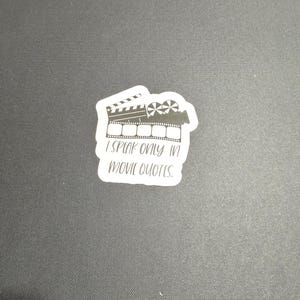 May include: White sticker with a black and white illustration of a clapperboard and film reel. The text "I speak only in movie quotes." is written below the illustration.