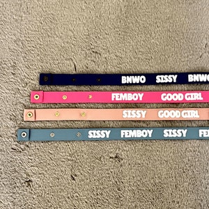 Könnte beinhalten: Vier farbige, schmale Lederarmbänder mit Text. Die Bänder sind marineblau, rosa, hellrosa und blaugrün. Der Text enthält "BNWO", "SISSY", "FEMBOY" und "GOOD GIRL". Jedes Band hat Druckknöpfe.