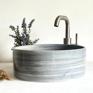 May include: A circular, grey and white ceramic basin with a brushed metal tap. The basin features a horizontal striped design. A small lavender plant is positioned to the left of the basin.