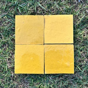 Könnte beinhalten: Vier quadratische, gelbe Fliesen in einem Raster angeordnet. Die Fliesen haben eine glatte, glänzende Oberfläche und sind vor einem Hintergrund aus grünem Gras angeordnet. Die Fliesen haben eine einfarbige, leuchtend gelbe Farbe.