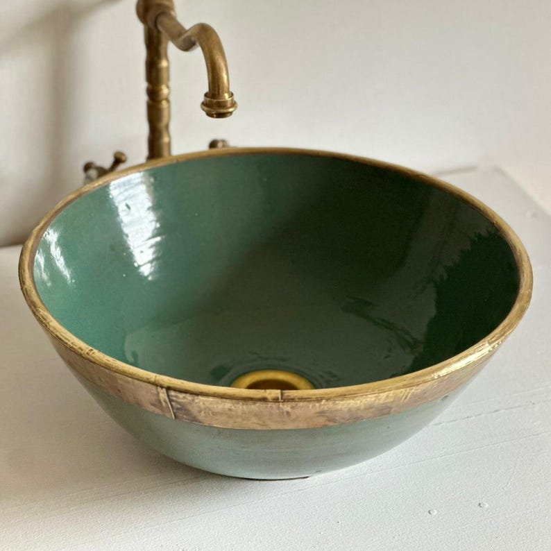 May include: A handcrafted, teal-colored ceramic vessel sink with a gold-toned rim. The sink has a round shape and a glossy finish. A vintage-style brass faucet is mounted above the sink, adding to its rustic charm. This sink is a unique addition to any bathroom.