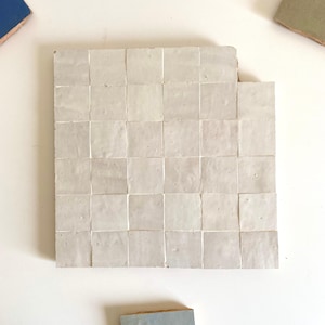 Puede incluir: Una disposición cuadrada de baldosas cuadradas de color blanco roto, posiblemente de cerámica o piedra, con líneas de lechada visibles. Las baldosas tienen una superficie ligeramente irregular, lo que sugiere una calidad artesanal o rústica. El color general es suave y neutro.