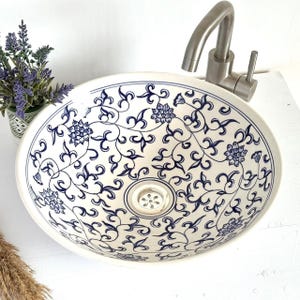 Może przedstawiać: Owalna ceramiczna umywalka z białą podstawą i niebieskim kwiatowym wzorem. Umywalka posiada kran i odpływ ze stali nierdzewnej. W tle widoczna jest mała roślina doniczkowa z fioletowymi kwiatami.