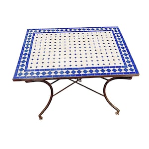 Peut inclure: Table de jardin carrée avec un dessus en mosaïque. Le plateau présente un fond blanc avec des carreaux bleus en forme de losange et carrés. La table a un cadre en métal foncé et des pieds incurvés. Mesure environ 76 cm x 76 cm.
