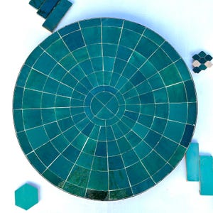 Puede incluir: Una tapa de mesa redonda de mosaico azul verdoso con un patrón circular de azulejos más pequeños. La tapa de la mesa está hecha de muchos azulejos pequeños y cuadrados que están dispuestos en un patrón circular. Los azulejos son todos de un tono azul verdoso y están separados por finas líneas de lechada blancas.