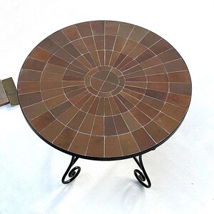 Peut inclure: Une table ronde en mosaïque brune avec un piétement en métal noir. Le plateau de la table est composé de petites tuiles carrées disposées en cercle.