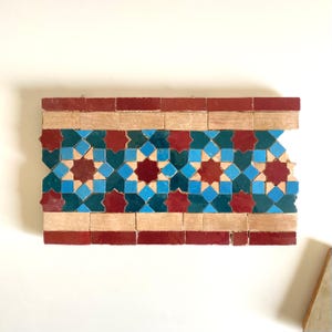 Peut inclure: Une bordure de carreaux rectangulaire avec un motif géométrique répétitif. Le motif présente des rangées alternées de carreaux rouges, beiges et turquoise. La rangée centrale présente des motifs en forme d'étoile et de losange en rouge, bleu et turquoise.