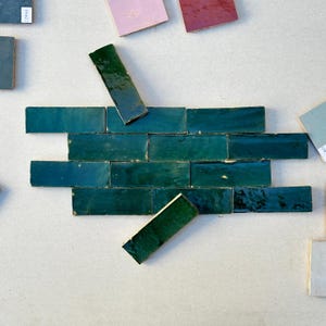 Op de afbeelding: Een presentatie van teal keramische tegels gerangschikt in een baksteenpatroon. Verschillende tegels zijn verspreid rond de hoofdaanrichting, met diverse kleurstalen op de achtergrond. De tegels hebben een glanzende afwerking.